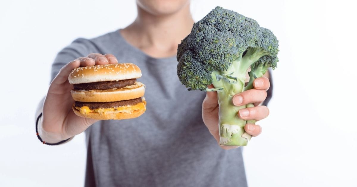 persona tiene in mano un hamburger e un broccolo per indicare come abbassare il colesterolo con la dieta