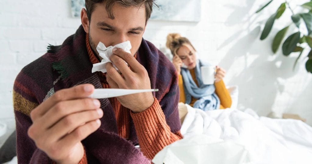 uomo e donna a letto, l'uomo ha il termometro e la donna si soffia il naso e si chiedono come combattere i malanni di stagione